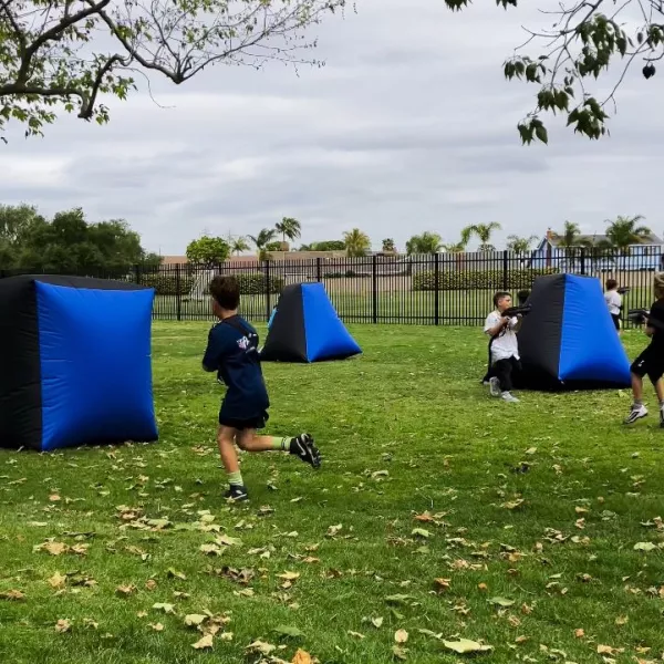 Kids playing laser tag between blue and black inflatable bunkers on a park lawn