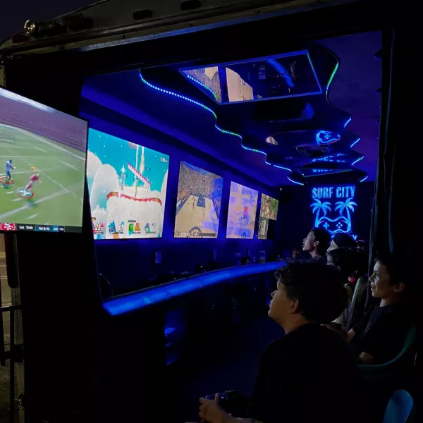 Inside the game truck — kids in stadium seating playing across a wall of screens under blue LED lighting