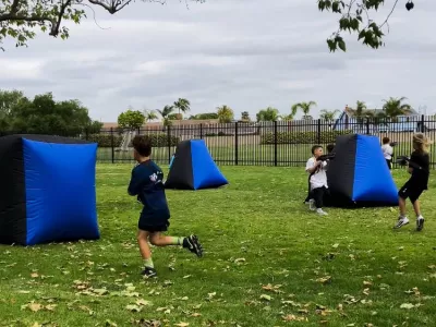 Kids playing laser tag between blue and black inflatable bunkers on a park lawn